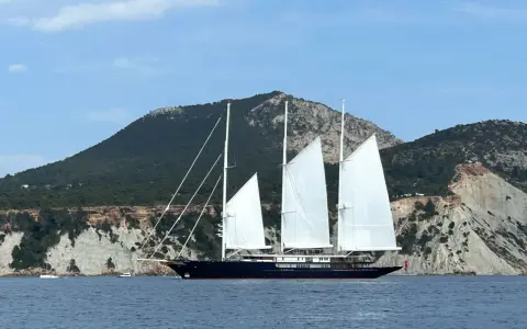 Le yacht de Bezos, un voilier majestueux, navigue en Méditerranée, ses voiles blanches déployées devant des falaises escarpées.
