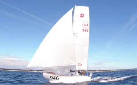 Un voilier de la classe mini, numéro 946, navigue sur une mer bleue sous un ciel clair.