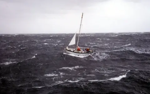 Un voilier blanc lutte contre les vagues déchaînées dans une mer agitée. Le ciel est gris et menaçant, annonçant une tempête.