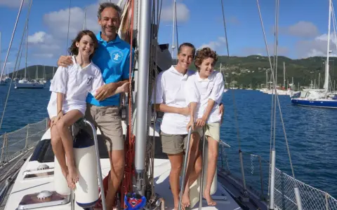 Famille souriante sur un voilier, un moment clé de leur récit de voyage en voilier. La mer bleue et les voiliers en arrière-plan complètent ce tableau idyllique.