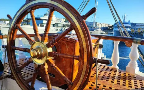 Roue de gouvernail en bois et laiton sur le pont d'un bateau. En arrière-plan, des yachts modernes et des bâtiments dans un port ensoleillé.