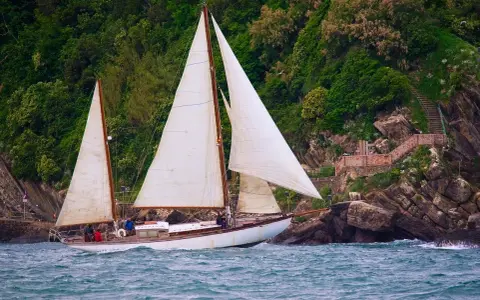 Un magnifique voilier 2 mâts navigue près d'une côte rocheuse verdoyante.
