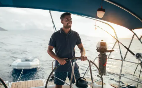 Un homme souriant barre un bateau, profitant du soleil couchant. Un petit bateau gonflable est attaché à l'arrière.