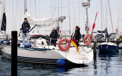 Trois personnes sur un bateau, prêtes à appareiller. Le drapeau allemand flotte fièrement. L'erre bateau est calme.