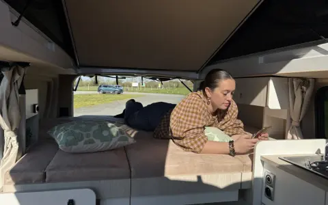 Une jeune femme se détend sur le lit de son camping-car j80 interieur, regardant son téléphone. Dehors, le soleil brille.