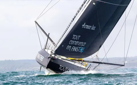 Le voilier IMOCA de Jean Le Cam fend les vagues, sa grand-voile noire ornée du slogan "Tout commence en Finistère" gonflée par le vent.