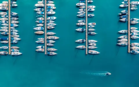 Vue aérienne d'un port où de nombreux voiliers sont amarrés. Un petit bateau navigue, laissant une trace dans l'eau. Le **bloc côtier obligatoire** est bien visible.