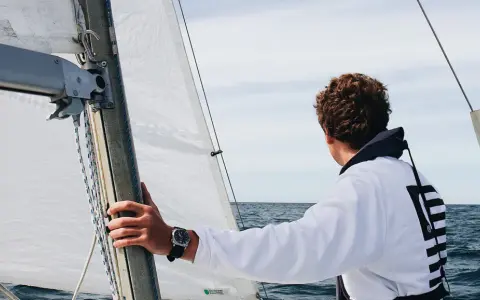 Un jeune homme regarde l'horizon depuis un voilier autonome. La voile blanche est gonflée par le vent.