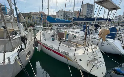 Un voilier First 32, amarré dans un port, avec d'autres bateaux et des bâtiments en arrière-plan.