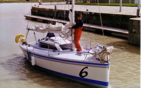 Un homme en salopette rouge prépare un voilier coco, le numéro 6 bien visible sur la proue, prêt à naviguer sur un canal.