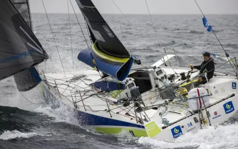 Un skipper navigue sur un voilier blanc et bleu, affrontant les vagues. Le nom "Pierre Quiroga" est visible sur la coque.