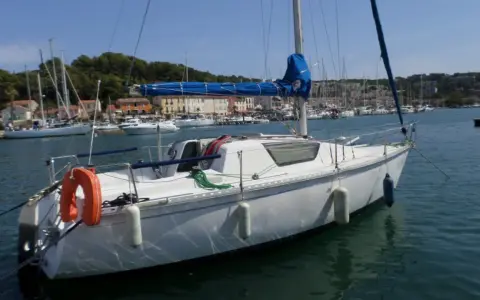 Bateau blanc à quai, voiles bleues enroulées. Au loin, un port animé avec des maisons colorées et d'autres voiliers. Un endroit idéal pour un séjour, comme le suggèrent les gib sea 76 avis.