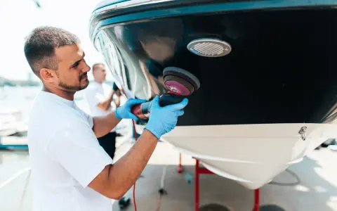 Un homme polit la coque d'un bateau. Il utilise une polisseuse pour un travail de précision, peut-être en prévision d'une révision moteur inboard.