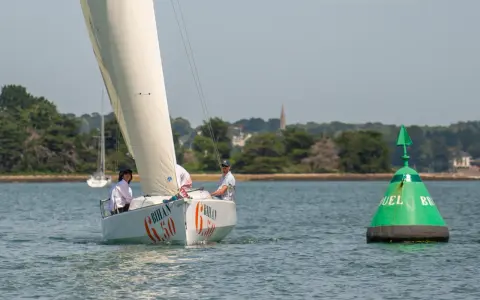 Voile blanche sur l'eau, le voilier "Bihan 6.50" navigue avec deux personnes à bord. Une bouée verte marque le passage.
