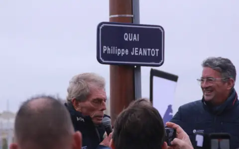 Un panneau "Quai Philippe JEANTOT" honore le navigateur. Philippe JEANTOT, entouré, parle au micro.