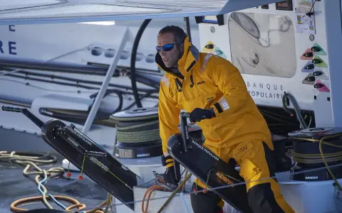 Skipper en tenue jaune, concentré, manœuvre un winch sur un bateau Banque Populaire.