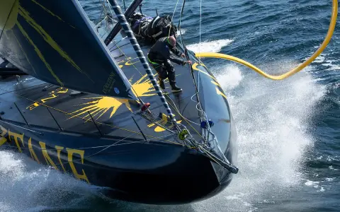 Un skipper manœuvre sur l'IMOCA Bureau Vallée, le bateau noir et jaune fend les vagues dans un embrun spectaculaire.