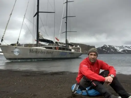 Mike Horn, l'explorateur, pose près de son bateau "Mercedes-Benz" sur une plage rocheuse, avec des montagnes enneigées en arrière-plan.