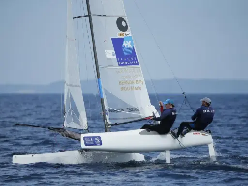 Deux marins, dont Billy Besson, naviguent sur un catamaran blanc par temps calme.