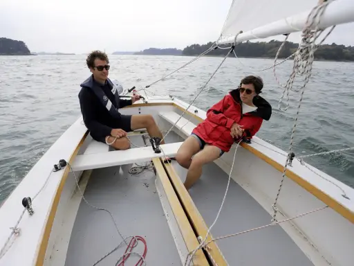Deux personnes profitent d'une journée ensoleillée sur un voilier dayboat, naviguant sur une mer calme.