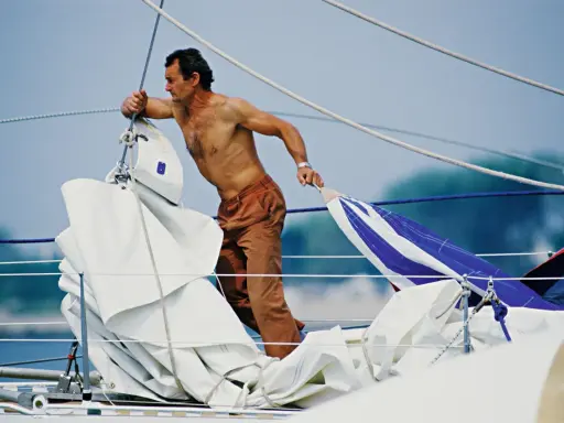 Un marinier, peut-être Eric Tabarly, manipule des voiles sur un bateau. Sa fortune est dans le vent et l'océan.