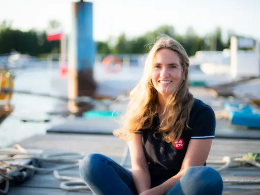 Irina Gracheva, souriante, assise sur un ponton en bois, avec des bateaux et des bouées en arrière-plan.