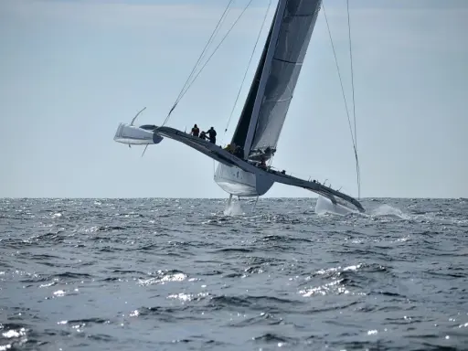Un maxi trimaran fend les vagues, ses coques latérales hors de l'eau, propulsé par une voile immense. L'équipage est visible sur le pont.