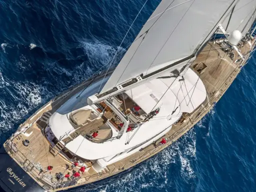 Le yacht "Bayesian" navigue sur des eaux bleues, ses voiles blanches gonflées par le vent. L'équipage en rouge s'affaire sur le pont en bois.