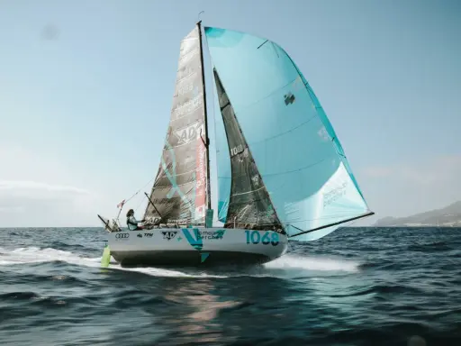 Un voilier de course, le numéro 1068, navigue sur une mer agitée. Les voiles, l'une noire et l'autre bleu clair, portent le nom "la maison perchée". Thais Le Cam est à la barre.