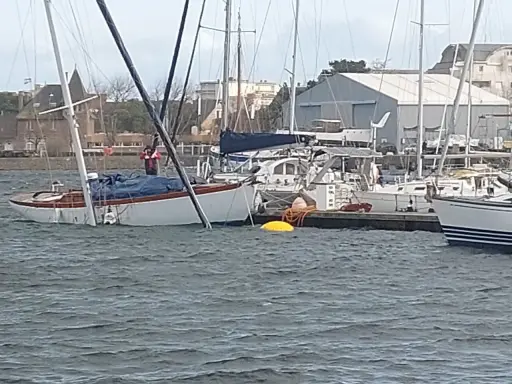 Un voilier blanc et bois, bâché, est partiellement immergé dans un port. Un homme en gilet rouge est à bord.