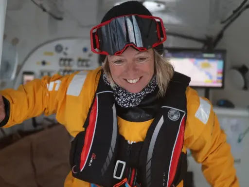 Alexia Barrier, souriante, porte une combinaison jaune et un gilet de sauvetage, prête pour une aventure maritime.
