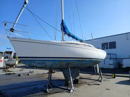 Voilier blanc sur bers, coque bleue usée, sous un ciel clair. Le mât est dressé, la grand-voile bleue enroulée. Premier 29 avis.