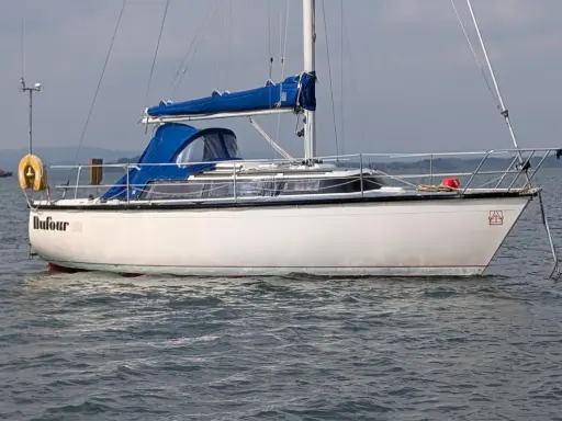 Voilier blanc Dufour 2800 à l'ancre, avec une bâche bleue sur la cabine. Un remorqueur orange est visible au loin.