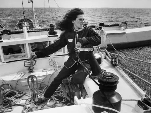 Une jeune femme aux cheveux longs, vêtue d'une combinaison de marin, manœuvre un winch sur un voilier. L'image évoque l'esprit d'aventure de Jacques Arthaud.