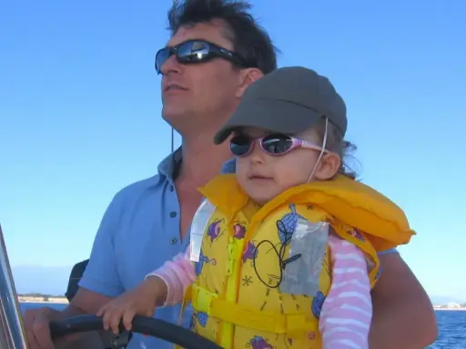 Un homme, le chef de bord bateau, tient son enfant sur ses genoux, naviguant sous un ciel bleu.