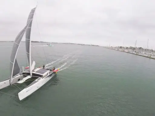 Un catamaran TS3 navigue sur une mer calme, avec un port rempli de voiliers à droite.