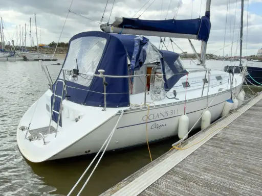 Un voilier blanc, un Océanis 311 Clipper, est amarré à un quai en bois. Des pare-battages blancs protègent sa coque.