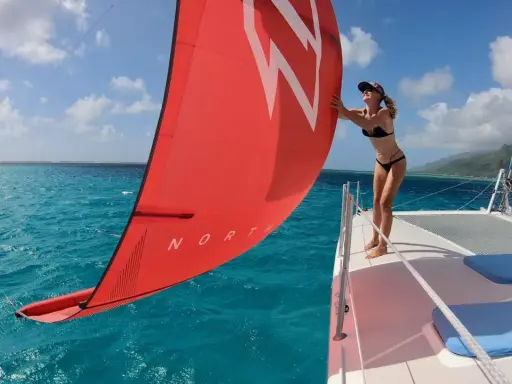 Une femme en bikini ajuste la voile rouge d'un petit catamaran habitable. L'eau turquoise et le ciel bleu créent un décor paradisiaque.