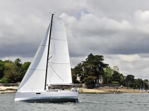 Voilier blanc moderne naviguant sur l'eau, avec des avis sur le Pogo 30. Paysage côtier verdoyant en arrière-plan.