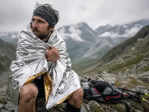 Un coureur transi, enveloppé dans une couverture de survie, contemple le paysage montagneux. Son sac à dos et ses bâtons de randonnée sont posés à côté.