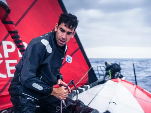 Gaston Morvan, marin concentré, ajuste une corde sur un voilier par temps couvert. La voile rouge porte des inscriptions blanches.
