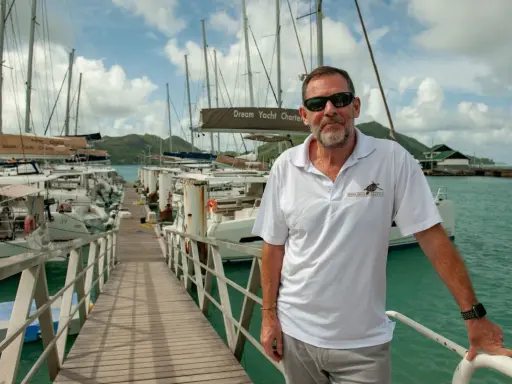 Loïc Bonnet, un homme souriant, pose sur un ponton bordé de voiliers. Il porte un polo Dream Yacht Charter, prêt pour une aventure en mer.