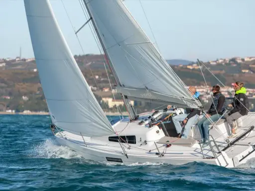 Un voilier First 27.7 navigue sur une mer agitée, avec quatre personnes à bord. Le soleil brille sur les voiles blanches et le paysage côtier en arrière-plan.
