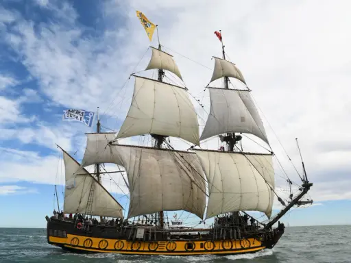 Le Shtandart, bateau historique aux voiles blanches, navigue sur une mer agitée sous un ciel nuageux.