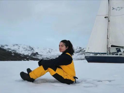 Tamara Klink, en tenue de ski jaune et noire, assise sur la neige, avec un voilier et des montagnes enneigées en arrière-plan.