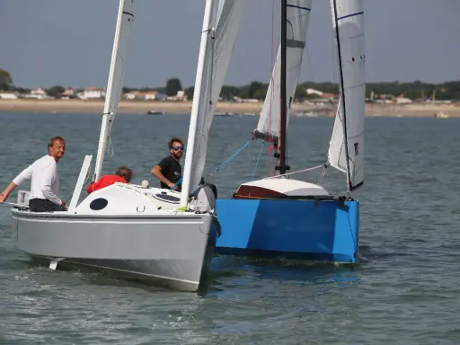 Deux voiliers naviguent côte à côte. Sur le premier, un homme en blanc manœuvre. Le second, d'un bleu vif, est un aloes 18.