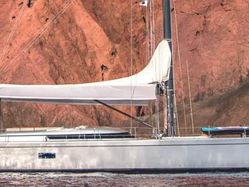 Un voilier Swan 76, avec une voile blanche déployée, navigue près d'une falaise rouge. Un kayak est rangé sur le pont.