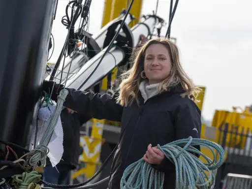 Élodie Bonafous, souriante, tient une corde bleue sur un bateau, entourée de cordages et de structures métalliques.