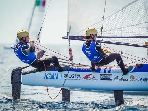 Deux jeunes navigateurs, dont Lou Berthomieu, en action sur un catamaran de sport, le vent gonflant les voiles.