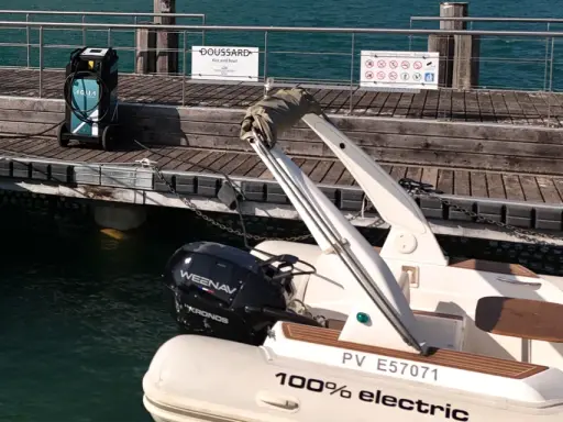 Un bateau électrique est prêt à être rechargé sur un ponton en bois. Une borne de recharge AQUA est visible à proximité.